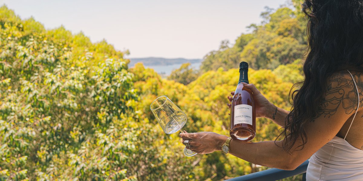 Alexandra Coccia holding a Champagne bottle and wine glass on a balcony overlooking the Central Coast